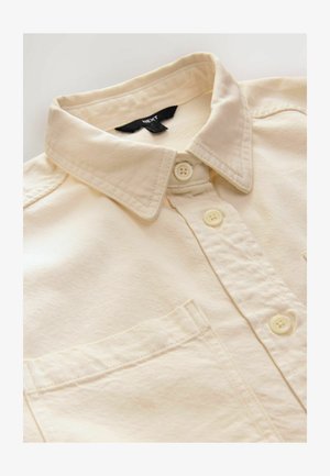 Kreminės spalvos mygtukų marškiniai su apykakle, turintys vieną krūtinės kišenę ir subtilią raištelių tekstūrą. Mygtukai yra kreminės spalvos ir apvalūs.