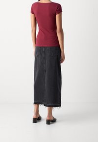 Femme portant un haut ajusté à manches courtes bordeaux, une longue jupe en jean noire à ourlet effiloché, et des chaussures noires à talons sans lacets, debout, de dos.