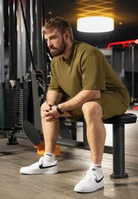 Completo sportivo verde oliva con t-shirt e pantaloni corti larghi. Sneakers bianche con accenti neri, seduto su una panchina in palestra. Ambiente fitness.