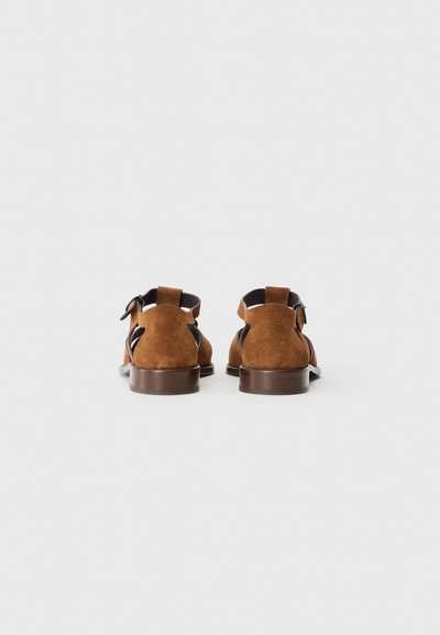 Sandales en suède marron avec dos ouvert, sangle réglable, accents noirs contrastés, petit talon marron et découpes sur les côtés.