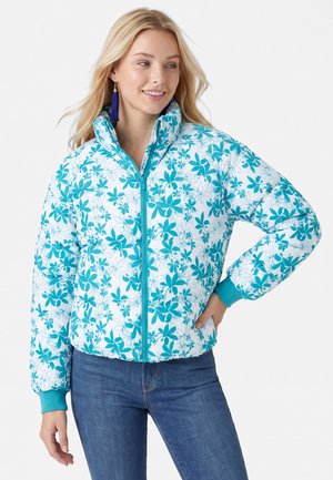 Frau trägt eine weiße Steppjacke mit türkisfarbenem Blumenmuster, türkisfarbenen Bündchen und Reißverschluss, kombiniert mit blauen Jeans und blauen Quastenohrringen.