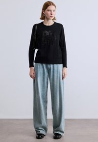 Maglione nero con logo "DKNY" borchiato, abbinato a pantaloni jeans larghi di colore azzurro chiaro e scarpe nere, con una borsa a tracolla.