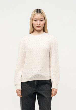 Vrouw met lang blond haar, gekleed in een wit gestructureerd gebreide trui en zwarte spijkerbroek, staand tegen een effen lichte achtergrond.