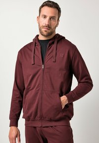 Sudadera con cremallera color marrón en tejido suave, con un bolsillo frontal, capucha ajustable con cordón y detalle del logo en el pecho.