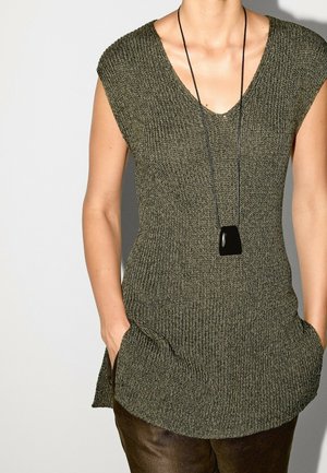 Femme portant un haut en tricot sans manches vert olive avec col en V et un collier pendentif noir, mains dans les poches, sur un fond uni.
