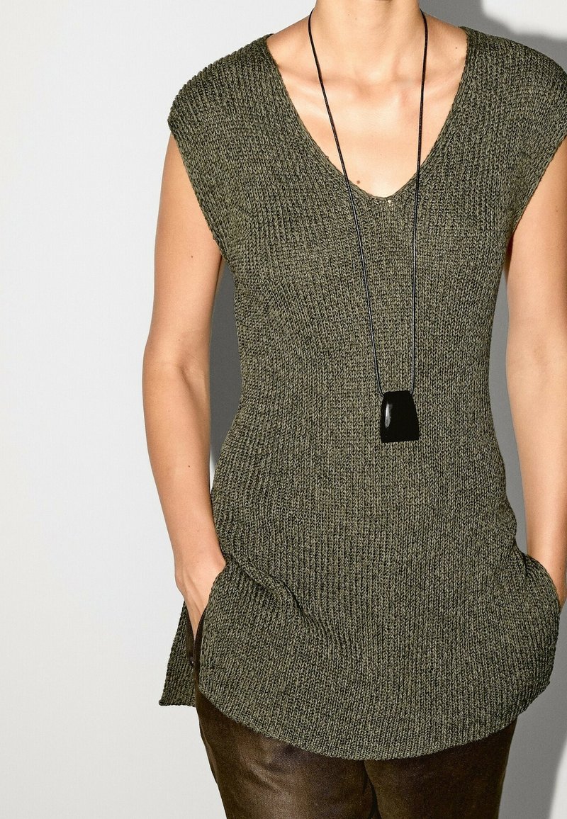 Femme portant un haut en tricot sans manches vert olive avec col en V et un collier pendentif noir, mains dans les poches, sur un fond uni.