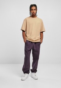 Beige übergroßes T-Shirt mit kurzen Ärmeln, kombiniert mit lockeren, lila Jogginghosen und weißen Turnschuhen. Neutraler Hintergrund.