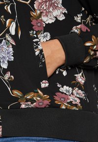 Svart tyg med blommönster har en ficka och ribbad mudd. Färgerna inkluderar rosa, vitt och grönt med en texturerad yta.