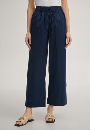 Frau trägt marineblaue Hosen mit weitem Bein und elastischem Bund sowie beige Pantoletten vor einem einfarbigen Hintergrund.