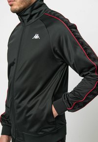 Zwarte trainingsjack gemaakt van een gladde stof, met een hoge kraag, ritssluiting aan de voorkant en rode accenten. Logo op de borst; patroon-details op de mouwen.