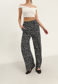 Pantalon noir à motif floral avec une coupe ample, une taille élastique et des poches latérales. Associé à un crop top blanc à épaules dénudées. Chaussures plates noires.