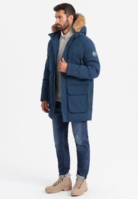  Marinblå parka med huva i fuskpäls, framdragkedja och tryckknappsstängning. Har två framfickor och en logotyp patch på ärmen. Bärs över en grå tröja.