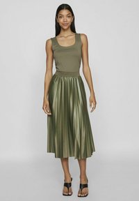 Falda plisada de color verde oliva hecha de tela brillante, combinada con una camiseta ajustada. El modelo lleva zapatos de tacón abiertos negros.