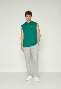 Grønn ermeløs hettegenser med snor og logo, kombinert med lysegrå joggebukser med en liten logodetalj, og hvite sneakers.