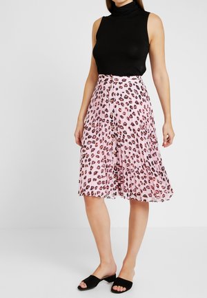 Femme portant un top noir sans manches à col roulé, une jupe midi plissée rose à imprimé léopard avec des boutons, et des sandales noires à talons ouverts à l’orteil.