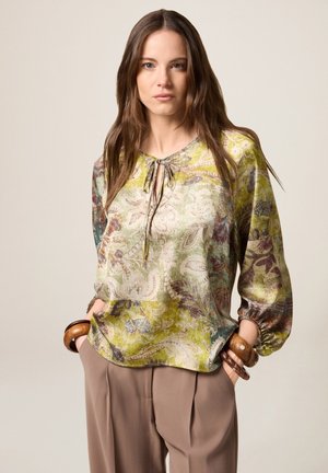 Silk blouse with a floral paisley pattern in green, purple, and beige. Features a tie neck and elasticized cuffs. Worn with beige trousers.