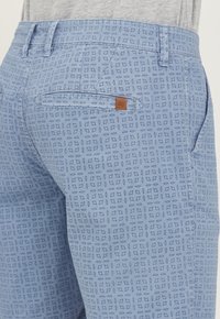 Pantaloni de culoare albastră deschisă cu un model geometric pe toată suprafața. Au o buzunar la spate cu o etichetă mică din piele maronie și un sistem de închidere cu fermoar.