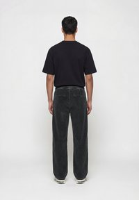 Άνδρας με κοντά μαύρα μαλλιά στέκεται γυρισμένος με την πλάτη, φοράει απλό μαύρο μπλουζάκι, σκούρα γκρι παντελόνια κοτλέ και λευκά αθλητικά παπούτσια.