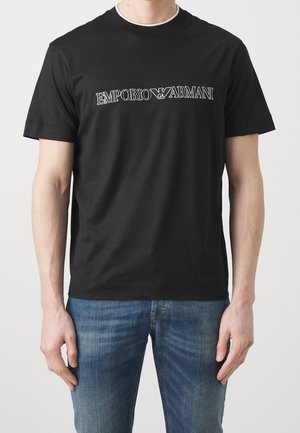 Mężczyzna w czarnej koszulce Emporio Armani z białym logo na piersi i niebieskich dżinsach, stojący na jednolitym tle.