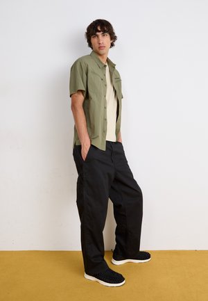 Ung mand står med hænderne i lommerne på sorte bukser, iført en olivengrøn kortærmet skjorte med knapper over en beige T-shirt og sorte sneakers.