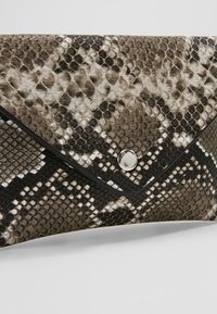 Pochette pliée avec motif peau de serpent marron, noir et blanc et fermeture par bouton argenté sur fond neutre.