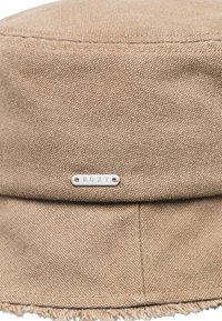 Brun stof bucket hat med tekstureret finish. Har et sølv "ROXY"-mærke og flossede kanter ved skyggen.