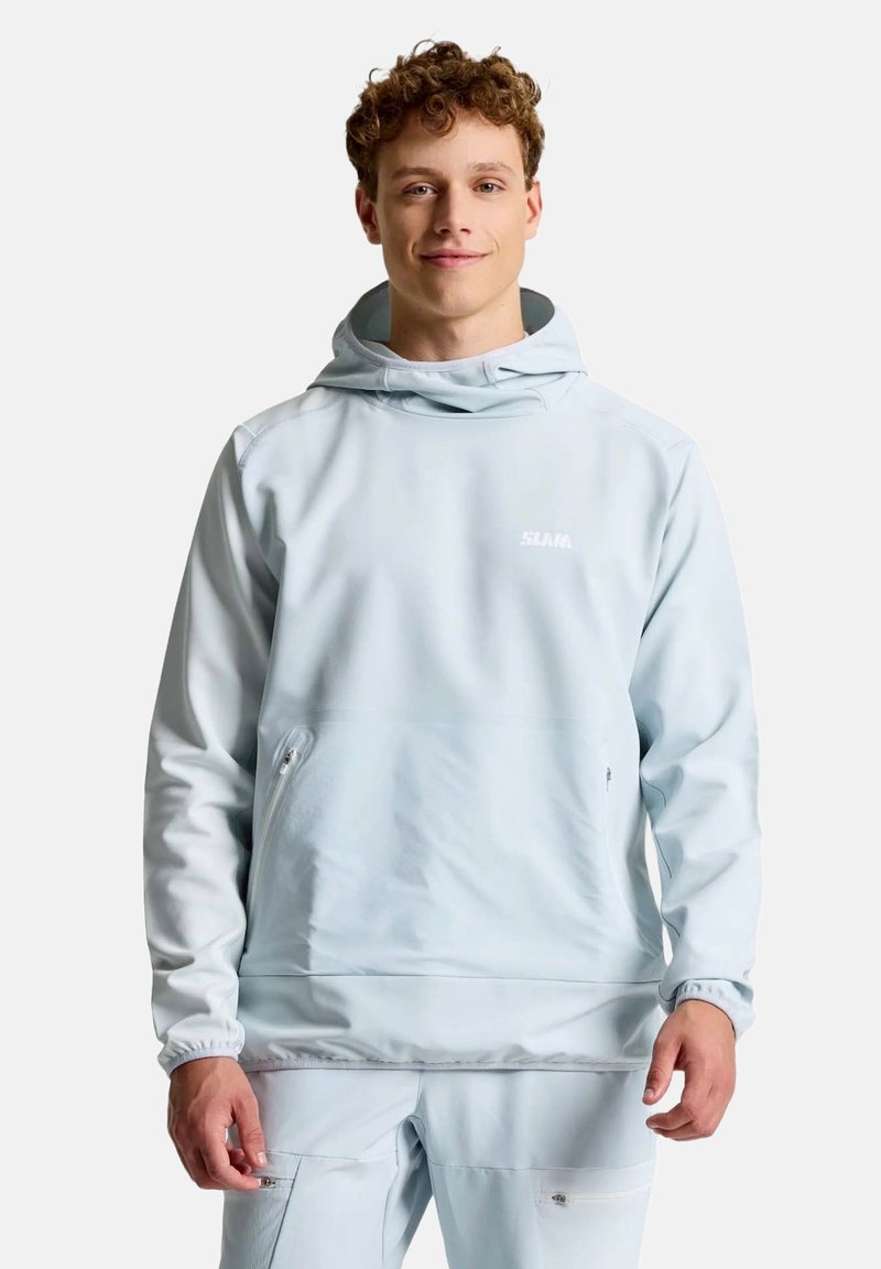 Jeune homme portant un sweat à capuche bleu clair et un pantalon assorti, debout et regardant vers l'avant avec un fond neutre.