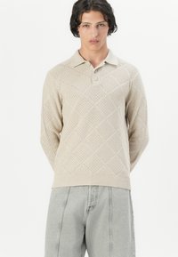 Sudadera de punto beige con un patrón de diamantes texturizado, que presenta un cuello con solapa y tres botones, combinada con pantalones gris claro.