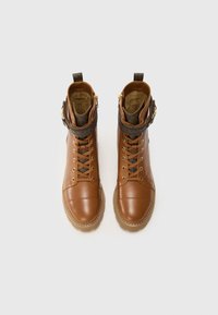 Bottes en cuir marron avec un bout rond, des lacets et une sangle décorative. Présentent un col texturé et des fermetures éclair sur le côté. Semelle en caoutchouc.