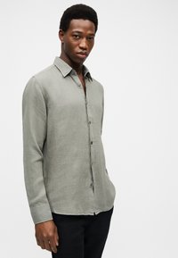 Νεαρός άνδρας στέκεται με τα χέρια στις τσέπες, φοράει ανοιχτό γκρι πουκάμισο με κουμπιά και μακριά μανίκια και μαύρο παντελόνι, μπροστά σε λευκό φόντο.