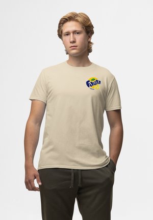 Ung mand iført en beige Fanta Lemon t-shirt og mørke bukser, stående med den ene hånd i lommen foran en ensfarvet baggrund.