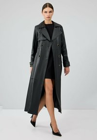 Lange zwarte leren trenchcoat met een dubbelzijdig ontwerp, grote kraag en gouden hardware. Heeft een hoge split en een strakke afwerking.