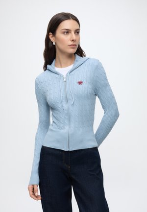 Femme aux cheveux foncés portant un sweat à capuche en maille torsadée bleu clair zippé avec un petit patch cœur rouge, un jean foncé et des boucles d'oreilles blanches, regardant vers la droite.