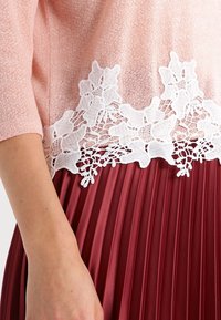 Femme portant un haut rose à manches courtes avec un bord en dentelle florale blanche sur une jupe plissée rouge, montrant le milieu du corps et le bras.