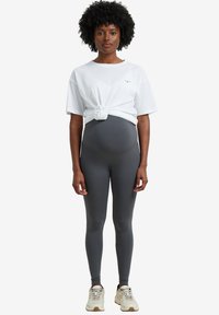 White knotted t-shirt over gray leggings, featuring a smooth texture and fitted design. Model wearing light-colored sneakers.