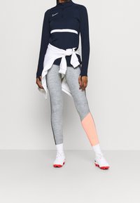 Camiseta deportiva de manga larga azul marino con una franja blanca; mallas grises con un detalle coral; zapatillas blancas con suelas rojas.