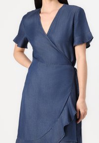 Denim wrap dress in dark blue, featuring short ruffled sleeves, a V-neckline, and a side tie closure. Flowy hem with a slight asymmetrical design.