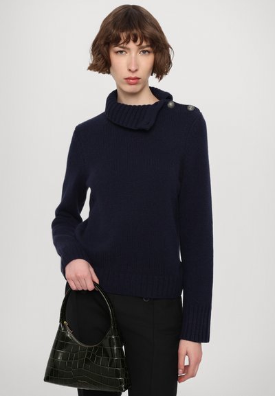 Pull en maille bleu marine avec un col roulé épais, présentant un détail de bouton sur une épaule. Le modèle tient un sac à main en cuir crocodile verni de couleur verte.