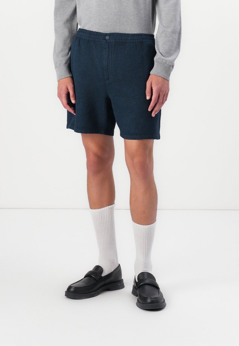 Dark blue, textured shorts with elastic waistband and button closure, paired with white crew socks and black slip-on shoes.