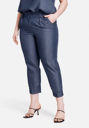 Vrouw draagt nauwsluitende donkerblauwe cropped broek met plooien, gecombineerd met zwarte sandalen met hak en bandjes, staand tegen een effen achtergrond.