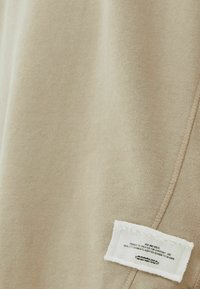 Detailní záběr na béžovou látku s bílou obdélníkovou štítkovou nášivkou, na které je malý černý tištěný text a viditelné stehy.