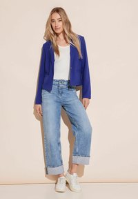 Krótkie niebieskie żakiet na białym T-shircie, zestawione z szerokimi, jasnoniebieskimi dżinsami. Białe sneakersy mają teksturowany wzór. Jasne tło.