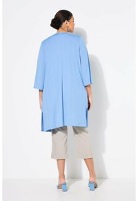 Helles blaues, langes Tunikaoberteil aus strukturiertem Stoff mit seitlichen Schlitzen, kombiniert mit beige farbenen, knielangen Hosen und hellblauen Sandalen mit Blockabsatz.