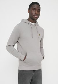 Jasnoszara bluza z kapturem wykonana z miękkiego materiału, z kieszenią kangurką i niewielkim haftowanym logo na lewej piersi. Klasyczny krój z regulowanym ściągaczem.