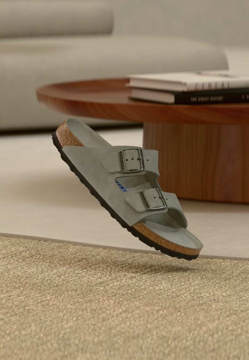 Γκρι σουέντ σανδάλι Birkenstock με δύο αγκράφες που αιωρείται πάνω από μπεζ χαλί κοντά σε ξύλινο τραπεζάκι καφέ με βιβλία.