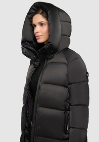 Jachetă de tip puf neagră cu glugă mare, prevăzută cu fermoar frontal, panouri cusute și buzunare laterale; textură netedă, lucioasă.