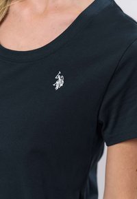 Temno mornarsko modra bombažna majica z okroglim izrezom in kratkimi rokavi. Na levi strani prsi majhen bel vezen logotip. Gladek tekstura.