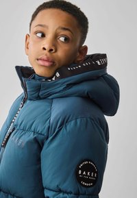 Doudoune en teal avec fermeture éclair sur le devant, grande capuche et accents noirs. Présente un patch logo sur la manche et un col intérieur rayé.