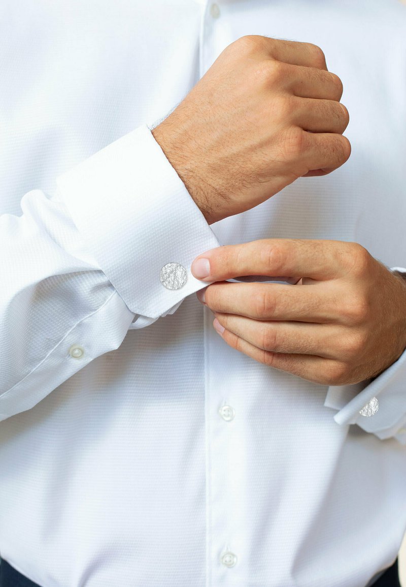 Camicia bianca con un motivo testurizzato, dotata di gemelli argentati sui polsini abbottonati. Mani che stanno aggiustando il polsino sinistro.