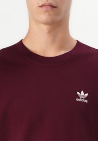 Burgundijas krāsas T-krekls ar apaļu kakla izgriezumu, izgatavots no kokvilnas, ar nelielu balto Adidas logo kreisajā krūšu daļā. Gluda tekstūra.